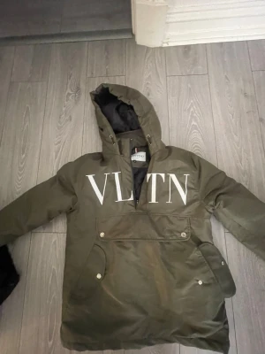 Olivgrön vindjacka från Valentino - Snygg olivgrön vindjacka från Valentino med stor vit VLTN-logga på bröstet. Jackan har huva, magficka med knappar och svart innerfoder. Perfekt för dig som vill ha en streetig och trendig look.