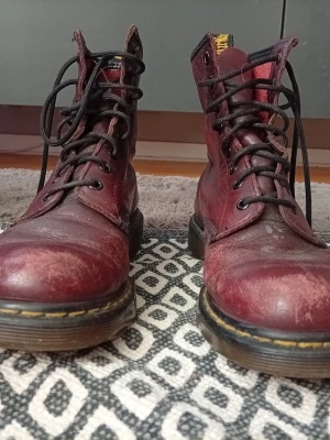 Vinröda Dr. Martens stövletter - Säljer ett par klassiska vinröda Dr. Martens stövletter i läder med svarta snören och grov sula. Ikonisk gul söm runt sulan och dragflik med logga bak. Perfekta för dig som vill ha en cool och tidlös look.