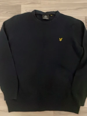 Svart tröja från Lyle & Scott - Klassisk svart tröja från Lyle & Scott med rund halsringning och den ikoniska gula logon broderad på bröstet. Tröjan har långa ärmar och ribbade muddar vid ärmslut och nederkant. Perfekt för en stilren och enkel look.
