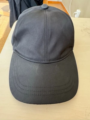 Svart keps från Prada - Snygg svart keps från Prada med klassisk böjd skärm och det ikoniska metalltriangelmärket på sidan. Tillverkad i slitstarkt tyg med stilren design och justerbar passform. Perfekt accessoar för en trendig och avslappnad look.