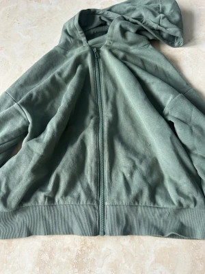 Grön zip hoodie med huva - En stilren grön hoodie med dragkedja framtill och huva. Tillverkad i mjukt bomullstyg med ribbade muddar vid ärmslut och nederkant. Perfekt för en avslappnad look och enkel att matcha med jeans eller mjukisbyxor.