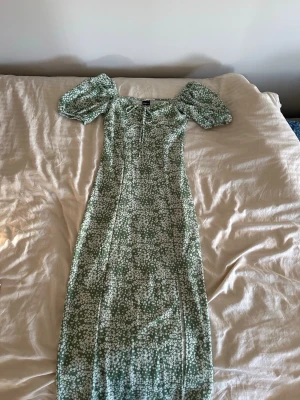 Grön blommig långklänning Gina Tricot - Supersöt grön långklänning från Gina Tricot med vitt blommönster, puffärmar och knyt framtill. Klänningen har en smickrande passform och är perfekt för vår och sommar. Materialet är mjukt och lätt, troligtvis bomull eller polyester.
