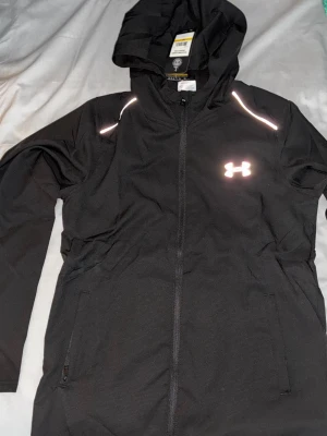 Svart windbreaker från Under Armour - Svart hoodie från Under Armour med dragkedja framtill och huva. Reflekterande detaljer på axlarna och logga på bröstet. Tillverkad i ett lätt och funktionellt syntetmaterial, perfekt för träning eller chill. Långärmad modell med fickor på sidorna.