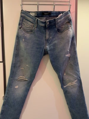 Replay anbass jeans - Säljer ett par ljusblå jeans från Replay med skinny fit och slitningar på både fram- och baksida. Jeansen har klassisk femficksmodell, dragkedjegylf och snygga detaljer som ger en trendig look. Perfekta för dig som gillar streetstyle.