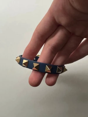 Valentino blått armband - Snyggt blått armband från Valentino i äkta läder med guldiga pyramidformade nitar runt om. Perfekt accessoar för dig som gillar en edgy och trendig stil. Armbandet har en smal design och justerbart spänne.