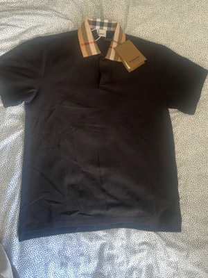 Svart Burberry t-shirt - Snygg svart t-shirt från Burberry med klassisk beige och röd-rutig krage. T-shirten har korta ärmar och en stilren passform. Perfekt för dig som vill ha en exklusiv och trendig look med ikonisk Burberry-detalj.