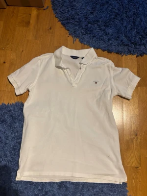 Vit piké från GANT med broderad logga - Klassisk vit pikétröja från GANT med korta ärmar och broderad logga på bröstet. Tröjan har krage och knappar framtill, tillverkad i mjuk bomull som känns skön mot huden. Perfekt för en clean och stilren look.