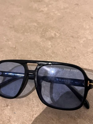Svarta Tom Ford pilotbågar solglasögon - Snygga solglasögon från Tom Ford med klassisk pilotform. Svart båge i plast och mörkblå tonade glas. Ikonis gulddetalj vid skalmen och diskret Tom Ford-logga på linsen. Perfekta för dig som vill ha en stilren och trendig look.