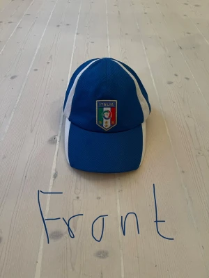 Blå Italia keps med broderad logga - Snygg blå keps med vita detaljer och broderad Italia-logga framtill. Justerbar rem baktill för perfekt passform. Tillverkad i ett lätt och ventilerande material, perfekt för att representera Italien på match eller i vardagen.