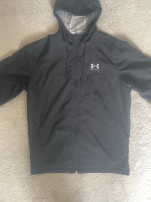Svart vindjacka från Under Armour - Snygg svart vindjacka från Under Armour med huva och dragsko. Jackan har meshfoder på insidan för extra ventilation och en vit logga på bröstet. Perfekt för blåsiga dagar och sportiga outfits.