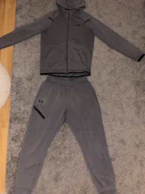 Grått loungewear-set från Under Armour - Byxorna är M och Koftan är S. En mysig grått loungewear-set från Under Armour med hoodie och matchande byxor. Hoodien har dragkedja och fickor, byxorna har resår i midjan och dragkedjeficka på sidan. Perfekt för chill dagar hemma eller när du vill vara bekväm.