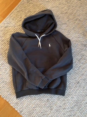 Extremt snygg hoodie från Polo Ralph Lauren - Mörkgrå hoodie från Polo Ralph Lauren med vit broderad logga på bröstet och vit snörning i huvan. Klassisk känguruficka framtill och ribbade muddar vid ärmslut och nederkant. Perfekt för en avslappnad och stilren look.