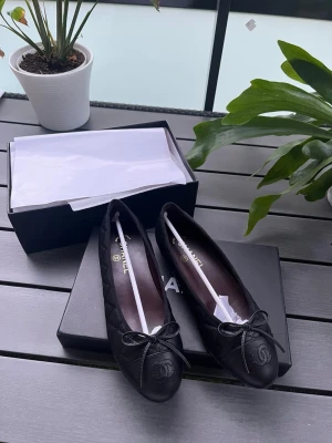 Svarta ballerinaskor från Chanel - Snygga svarta ballerinaskor från Chanel i quiltat läder med rund tå och dekorativ rosett framtill. Klassisk design med diskret CC-logo på tån. Perfekta för en stilren och lyxig look.