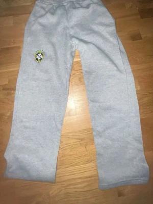 Grått Brasilien mysdress-set - Säljer ett grått mysdress-set med Brasilien-logga på både byxan och hoodien. Hoodien har huva och magficka, byxorna har resår i midjan. Perfekt för chill dagar eller när du vill visa support för Brasilien. Mjuk och skön bomullsblandning.