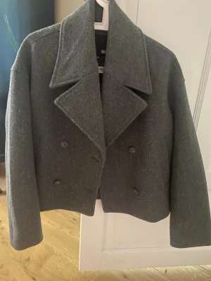Grå skön kappa  - Snygg grå kappa från lager 157 med bred krage och dubbelknäppning framtill. Kortare modell som ger en modern look. Perfekt till hösten och vintern. Klassisk design med raka ärmar och mjukt ullmaterial.