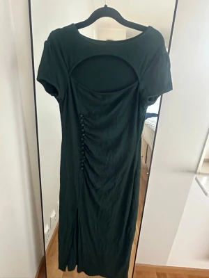 Mörkgrön långklänning med cut out - Snygg mörkgrön långklänning med korta ärmar och cut out-detalj i ryggen. Klänningen har dekorativa knappar längs sidan och en liten slits nedtill. Tillverkad i mjukt tyg som ger en smickrande passform.