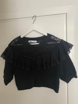Kofta/topp med volang  - Snygg svart kofta/topp från Zara med dekorativa virkade detaljer och dubbla volanger över axlar och bröst. Koftan har knäppning framtill och halvlånga ärmar. 