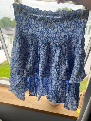 Blå blommig volangkjol - Supersöt kort kjol med blått och vitt blommönster och volanger. Kjolen har smock i midjan för en bekväm passform och är perfekt för varma dagar. Tillverkad i ett lätt och luftigt material som känns skönt mot huden.