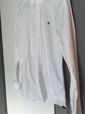 Vit skjorta från Morris Stockholm - Klassisk vit skjorta från Morris Stockholm med button down-krage och liten broderad logga på bröstet. Skjortan är tillverkad i bomull och har långa ärmar samt knappar framtill. Perfekt för en clean och stilren look.