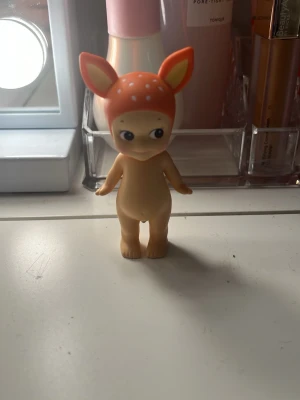 Sonny angel - En liten samlarfigur i plast med beige kropp och ett hjorthuvud i orange med vita prickar och stora öron. Perfekt som dekoration eller leksak för den som gillar gulliga djurmotiv.