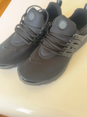 Svarta Nike presto - Snygga helsvarta Nike sneakers med ovandel i mesh och detaljer i plast vid snörningen. Skorna har rund tå, tjock sula och klassisk Nike-logga på sidan och tungan. Perfekta för dig som gillar en clean och sportig look.