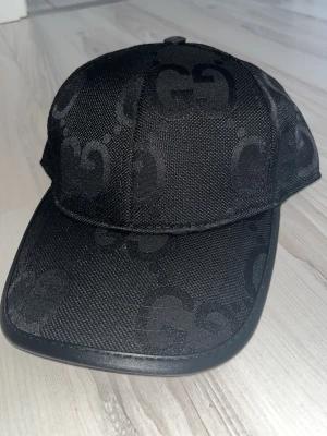 Svart keps med GG-mönster från Zara - Säljer en svart keps från Zara  med det klassiska GG-mönstret ton-i-ton över hela kepsen. Kepsen har böjd skärm och är tillverkad i ett slitstarkt tygmaterial. Perfekt för dig som vill ha en stilren och exklusiv accessoar.