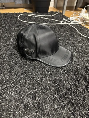 Svart keps från Prada i nylon - Snygg svart keps från Prada i glansigt nylonmaterial med klassisk form och böjd skärm. Prada-loggan sitter på sidan i metall. Perfekt för dig som vill ha en stilren och exklusiv accessoar till din outfit.