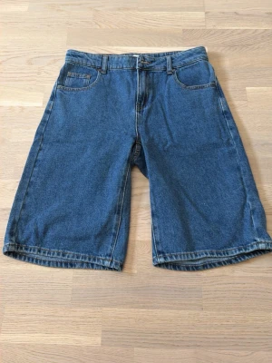 Blå denimshorts i loose fit från Grunt  - Säljer ett par klassiska blå jeansshorts i loose fit med fem fickor och knappgylf. Perfekta för dig som gillar avslappnad stil och vill ha gott om rörelseutrymme. Tidlös modell som passar till det mesta.