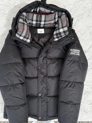 Svart pufferjacka från Burberry - Säljer en svart pufferjacka från Burberry med huva och rutigt innerfoder i grått, vitt och rött. Jackan har tryckknappar och dragkedja framtill samt logga på ärmen. Perfekt för kalla vinterdagar och riktigt snygg streetstil. Man kan även ta bort armarna så det blir en väst. Priset är inte hugget i sten vid snabb affär. Hör av er vid minsta lilla tillfälle MVH Filip.