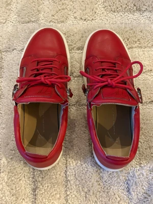 Röda sneakers från Giuseppe Zanotti - Snygga röda sneakers i läder från Giuseppe Zanotti med vita sulor, röda skosnören och silverfärgade dragkedjor på sidorna. Skorna har en låg profil och coola detaljer som ger en unik look. Perfekta för dig som vill sticka ut med din stil.