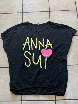 Svart Anna Sui t-shirt med tryck storlek XL - Cool svart t-shirt från Anna Sui med stort gult tryck och ett rosa hjärta på framsidan. T-shirten har korta ärmar och en avslappnad passform. Perfekt för dig som vill sticka ut med en unik och trendig design.