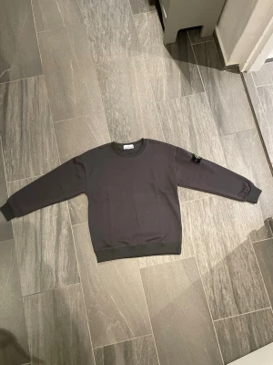 Mörkgrå sweatshirt från Stone Island - Säljer en stilren mörkgrå sweatshirt från Stone Island med den klassiska logotypen på vänster ärm. Tröjan har rund halsringning, ribbade muddar och är tillverkad i mjukt bomullsmaterial. Perfekt för dig som gillar streetwear och vill ha något enkelt men snyggt.