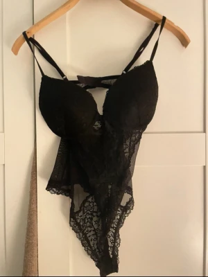 Svart spetsbody med tunna axelband - Snygg svart body i spets med tunna justerbara axelband och vadderade kupor. Djup urringning och transparent spetsdetalj framtill ger en sexig look. Perfekt för dig som vill ha något extra under kläderna.
