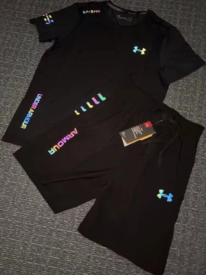Svart träningsset från Under Armour - Svart träningsset från Under Armour med färgglada loggor och textdetaljer i regnbågsfärger. Klassisk passform med snörning i midjan och tillverkade i ett mjukt och funktionellt material som passar perfekt till träning. Ikonisk UA-logga på ena benet. Storlek S, helt ny 