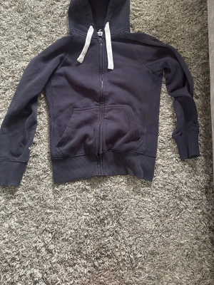 Mörkblå russemerch hoodie - Mörkblå hoodie med vit dragsko och dragkedja framtill. Klassisk känguruficka och ribbade muddar vid ärmslut och nederkant. Inte alls sliten 