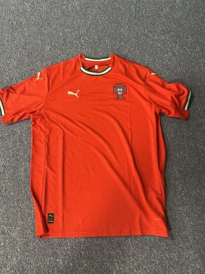 Röd Portugal fotbollströja Puma - Snygg röd fotbollströja från Puma med Portugals landslagsemblem på bröstet. Tröjan har vita och gröna detaljer vid krage och ärmslut samt Puma-logga på bröst och ärm. Tillverkad i lätt och ventilerande material, perfekt för träning eller match.