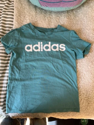Turkos Adidas t-shirt med logga - Snygg turkos t-shirt från Adidas med stor vit logga framtill. Klassisk passform och rund halsringning. Perfekt för en sportig och avslappnad stil. 