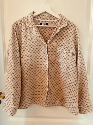 Pyjamaströja DKNY - Snygg beige pyjamaströja från DKNY med svart grafiskt mönster och vita kontrastkanter. Klassisk krage, knappstängning framtill och en bröstficka. Perfekt för mysiga kvällar hemma.