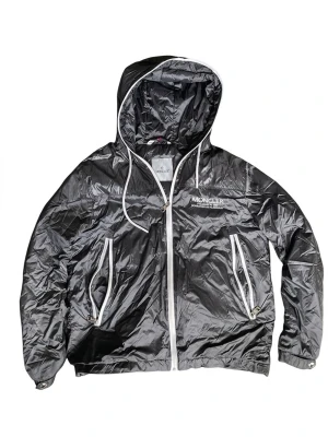 Grå vindjacka från Moncler - Snygg grå vindjacka från Moncler med vit dragkedja och vita detaljer. Jackan har huva med dragsko, två fickor med dragkedja och Moncler-logga på ärmen. Tillverkad i glansigt, lätt material som ger en sportig look. Finns en liten skada på den vänstra armen som kan fixas.