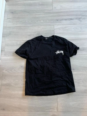 Svart Stüssy t-shirt med logga - Svart t-shirt från Stüssy med klassisk vit logga tryckt på bröstet. T-shirten har rund halsringning och korta ärmar. Tillverkad i mjuk bomull för en skön och avslappnad stil.