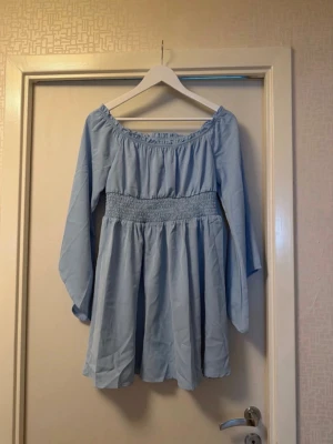 Ljusblå offshoulder klänning - Söt ljusblå kortklänning med offshoulder-design och långa ärmar. Klänningen har smock i midjan som ger en fin passform och ett luftigt fall. Perfekt för varma dagar och enkel att styla med accessoarer.