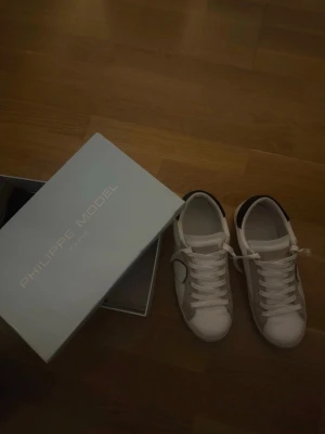 Vita Philippe Model sneakers - Snygga vita sneakers från Philippe Model med klassisk låg silhuett. Skorna har vita skosnören, detaljer i beige mocka och svart hälparti. Ikonisk sköldlogga på sidan och stilren design som passar till det mesta.