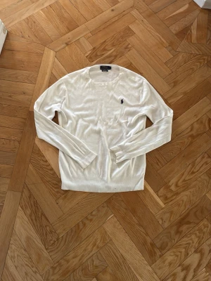Vit stickad tröja från Polo Ralph Lauren - Klassisk vit stickad tröja från Polo Ralph Lauren i slim fit-modell. Tillverkad i mjuk pima bomull och har den ikoniska broderade loggan på bröstet. Långärmad med rund hals och ribbade muddar för en clean look.