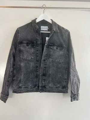 Grå jeansjacka Calvin Klein Jeans - Helt oanvänd grå jeansjacka från Calvin Klein Jeans med broderad CK-logga och text på ryggen. Klassisk modell med två bröstfickor med lock och knappar framtill. Storlek: 164 cm. Nypris/ 1099 kr