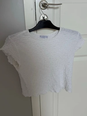 Vit croppad topp från Stradivarius - Säljer en vit croppad topp från Stradivarius med korta ärmar och ribbad struktur. Toppen har en lätt vågig kant nertill och är perfekt för en avslappnad stil. Enkel att matcha med jeans eller kjol.