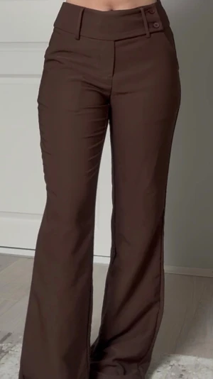 Bruna utsvängda kostymbyxor - Snygga bruna kostymbyxor med hög midja och utsvängda ben. Byxorna har två dekorativa knappar i midjan och en stilren design som ger en modern siluett. Perfekta för dig som gillar trendiga och klassiska plagg i garderoben. Har använt dem endast en gång. 