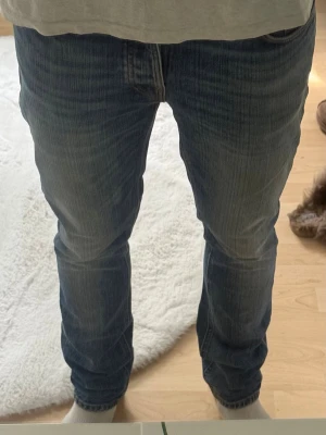 Blå raka jeans från Nudie Jeans - Säljer ett par klassiska blå jeans från Nudie Jeans med rak passform och snygga slitningar. Jeansen har fem fickor, normal midja och är gjorda i mjuk denim. Perfekta för en avslappnad och stilren look.