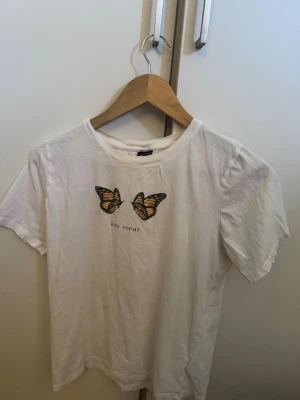Vit t-shirt med fjärilar och text - Vit t-shirt i bomull med tryck av två orangea fjärilar och texten 'âme soeur' på bröstet. Klassisk rund halsringning och korta ärmar. Perfekt för en chill och stilren look.