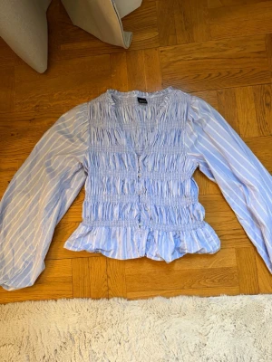 Blå- och vitrandig blus med volang - Söt blus i ljusblått med vita ränder, smockad överdel och volangkant nedtill. Blusen har knappar framtill och långa puffiga ärmar. Perfekt för en trendig och avslappnad look. Knappt använd
