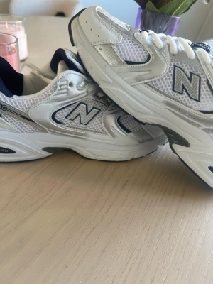 New Balance 530  - Snygga New Balance 530 sneakers i vitt med silver- och marinblå detaljer. Ovandel i mesh och syntet för en sportig look, med ABZORB-dämpning i sulan för extra komfort. Klassisk låg modell med tydlig N-logga på sidan.